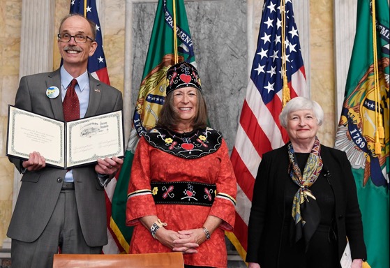 Director Len Olijar, Treasurer Lynn Malerba, and Secretary Janet Yellen standing together in front of American and Treasury Flags. Len Olijar is holding a document showing Lynn Malerba's signature.